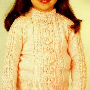 Puede incluir: Un suéter de punto rosa pálido con un patrón de punto de cable y un cuello alto. El suéter tiene un diseño decorativo de hojas y flores tejidas.