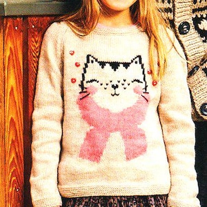 May include: A young person wearing a light beige sweater with a cat face design and a pink scarf. The sweater has red heart accents. They are also wearing a pink knit hat and a patterned skirt. The background includes a wooden wall.