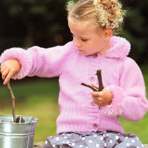 May include: A young girl wearing a pink knitted cardigan with fuzzy trim on the cuffs and collar. She is holding two sticks in her hands and is looking down at them.