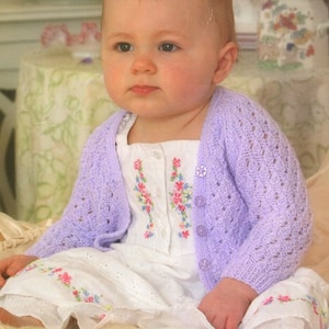 Puede incluir: Un bebé con un cárdigan de punto lavanda y un vestido blanco con bordado floral. El cárdigan tiene cierre de botón y un delicado patrón de calado.