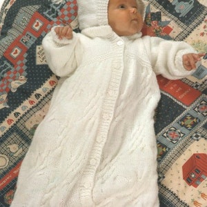 May include: A white knitted baby sleeper with a hood and button closure. The sleeper has a cable knit pattern and is lying on a colorful patchwork quilt.