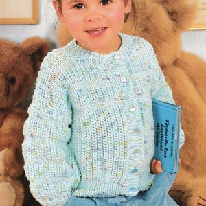 May include: A light blue knit cardigan with button closures. The cardigan has a speckled pattern of various colors. The child is holding a blue book titled "Thomas the Tank Engine and the Tractor".