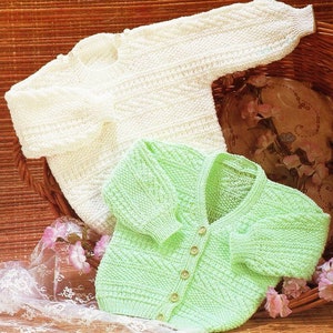 Puede incluir: Dos jerséis de punto para bebé, uno blanco y otro verde claro, con botones. Los jerséis se muestran en una cesta de mimbre.