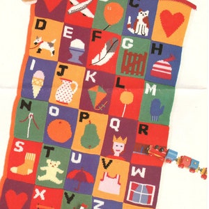 Könnte beinhalten: Eine bunte gestrickte Alphabet-Decke mit verschiedenen Bildern für jeden Buchstaben. Die Decke besteht aus quadratischen Feldern in verschiedenen Farben und zeigt ein rotes Herz, ein Flugzeug, einen Ballon, eine Katze, einen Hund, ein Ei, eine Feder, einen Zaun, einen Hut, ein Eis am Stiel, eine Kanne, einen Drachen, einen Lolli, einen Fäustling, eine Nadel, eine Orange, eine Birne, eine Königin, einen Zug, eine Socke, einen Teddybär, einen Regenschirm, ein Kleid, ein Fenster, eine Leiter, ein Segelboot und einen Reißverschluss.
