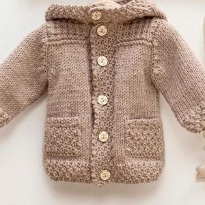 May include: A brown knitted baby cardigan with a hood and button closure. The cardigan has two pockets on the front.