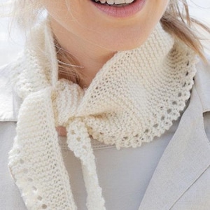 May include: A white crocheted scarf with a delicate lace trim. The scarf is tied around the neck of a person wearing a beige jacket.