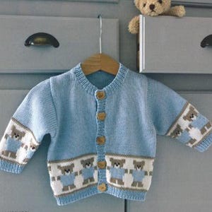 Może przedstawiać: Jas dla niemowlęcia w jasnoniebieskim kolorze, wykonany z dzianiny, z wzorem brązowych misiów i drewnianymi guzikami. Jas jest zawieszony na drewnianym wieszaku.