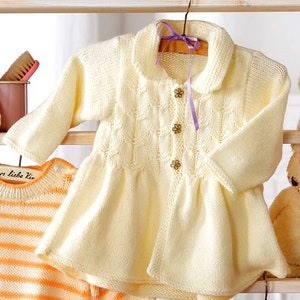 May include: A white knitted baby cardigan with a ruffled hem and a collar. The cardigan has a button closure and a purple ribbon tied around the collar.