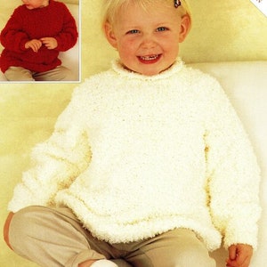 May include: A young child wearing a white knitted sweater with a high neck. The sweater has a fluffy texture. The child is sitting on a light brown surface and wearing beige pants and red shoes.