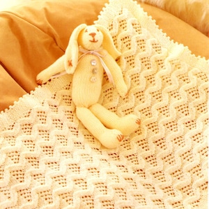 May include: A handmade, light yellow knitted bunny doll with long ears, a pink ribbon, and button details, resting on a cream-colored, patterned knitted blanket. The blanket has a textured, wave-like design.