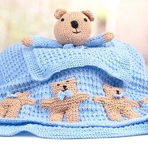 Puede incluir: Una manta de bebé de punto azul con un oso de peluche marrón y tres ositos más pequeños unidos. La manta tiene pompones azules en los bordes.