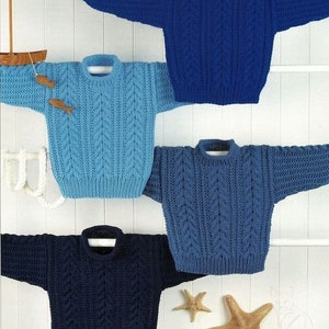 Könnte beinhalten: Vier gestrickte Pullover in verschiedenen Blautönen, die an einer weißen Wand hängen. Die Pullover haben ein Zopfmuster und sind alle im gleichen Stil.