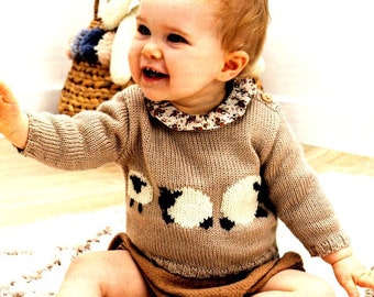 May include: A baby wearing a beige knitted jumper with a white and black sheep design. The jumper has a frilled collar with a floral pattern.
