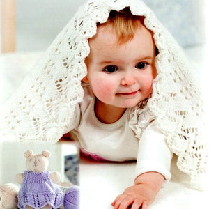 Può includere: Una copertina per neonati in crochet bianco con un delicato motivo a pizzo copre un bambino sdraiato su una superficie bianca. Il bambino indossa un body bianco e ha un orsacchiotto di peluche lavorato a maglia in primo piano.