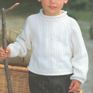 May include: A white knitted sweater with a ribbed texture and a crew neck. The sweater has long sleeves and is worn with dark blue jeans. The child is holding a wooden stick.