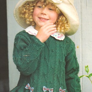 May include: A young girl wearing a green knitted sweater with a white collar and a white hat with white flowers. The sweater has a ruffled hem and three bows on the front. The girl is smiling and holding a basket.