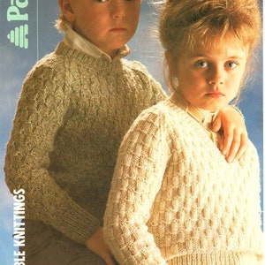 May include: Two children wearing knitted sweaters. The boy is wearing a light green sweater with a cable knit pattern. The girl is wearing a cream colored sweater with a textured knit pattern. Both sweaters have a V-neck.