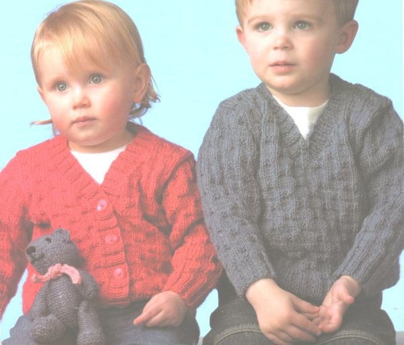 May include: Two children wearing knitted cardigans. The girl is wearing a red cardigan with buttons and a teddy bear. The boy is wearing a gray cardigan.