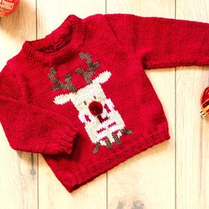 May include: A red knitted sweater featuring a reindeer design on the front. The reindeer has a white face, brown antlers, and a red pom-pom nose. The sweater is displayed on a wooden surface with Christmas ornaments.
