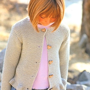 May include: A light gray knitted cardigan with a button-up front and wooden heart-shaped buttons. The cardigan has a slightly ruffled hem and is worn over a pink t-shirt.