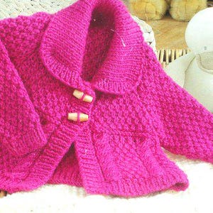 May include: A vibrant pink knitted baby jacket with a rounded collar and wooden toggle closures. The jacket is displayed on a woven surface, with a stuffed duck toy in the background. The jacket has a textured knit pattern.