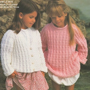 Könnte beinhalten: Zwei Mädchen tragen gestrickte Pullover. Das Mädchen links trägt eine weiße Strickjacke mit Perlmuttknöpfen und einen rosa Blumenrock. Das Mädchen rechts trägt einen rosa Pullover. PBN D 2078 Für die Größen 2-11 Jahre 56-76 cm