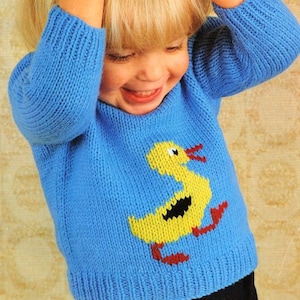 May include: A blue knitted sweater with a yellow duck applique. The duck has black eyes and a black beak.