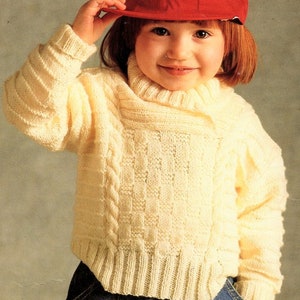 May include: A cream-colored, knitted sweater with a cable knit design on the front and ribbed cuffs. The sweater has a turtleneck and is worn with a red baseball cap and blue jeans. The sweater is a cozy and warm garment.