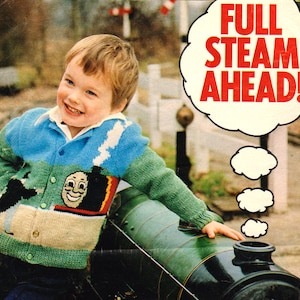 Pode incluir: Um menino pequeno vestindo um casaco de tricô verde, azul e branco com um motor de trem de desenho animado na frente. O casaco tem fecho de botão. O menino está de pé ao lado de um motor de trem de brinquedo verde. Uma bolha de fala acima da cabeça do menino diz "FULL STEAM AHEAD!"