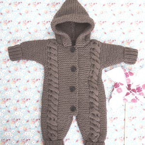 May include: A brown knitted baby jumpsuit with a hood. The jumpsuit features a cable knit design on the front and sleeves, with three dark buttons down the center. The hooded jumpsuit is designed for a baby or toddler.