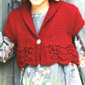 Könnte beinhalten: Ein roter Strick-Bolero mit Schalkragen und einem einzelnen Knopfverschluss. Der untere Rand hat ein dekoratives Lochmuster. Der Bolero wird über einem Kleid mit Blumenmuster getragen.