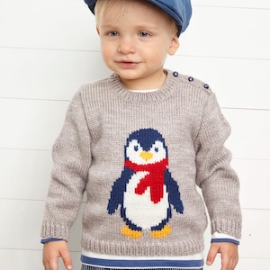 May include: A light brown knitted sweater with a penguin design, featuring a red scarf, blue wings, and yellow feet. The sweater has a crew neck and blue buttons on the shoulder. The child is wearing a blue cap.