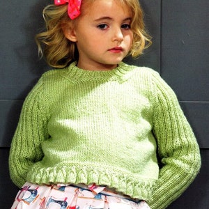 May include: A young girl wearing a light green knitted sweater with a ruffled hem and a pink bow in her hair. She is wearing a white skirt with a colorful sewing machine pattern.