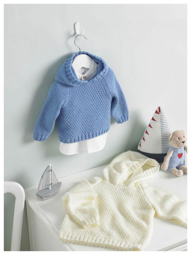 May include: A light blue knitted baby hoodie with a white collared shirt underneath, hanging on a white hanger. A second cream-colored hoodie is on a white surface, along with a toy sailboat and a stuffed animal.