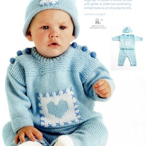 May include: A baby wearing a light blue knitted sweater and pants set with a white heart design. The set includes a matching hat with a white heart design. The sweater has knitted buttons on the shoulders.