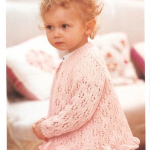 May include: A pink knitted cardigan with a delicate lace pattern and a ruffled hem. The cardigan is worn by a young child with curly blonde hair.
