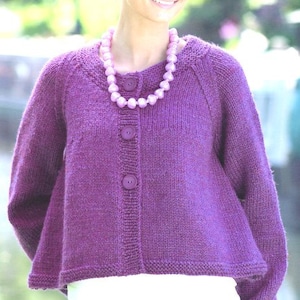 Puede incluir: Un cárdigan de punto morado con tres botones grandes y mangas largas. El cárdigan se combina con una camiseta blanca y un collar de cuentas. El cárdigan es de color liso y tiene un aspecto texturizado.