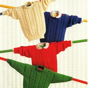 Op de afbeelding: Vier gebreide truien in verschillende kleuren hangen aan een waslijn. De truien zijn crème, blauw, rood en groen. Elke trui heeft een ander patroon en is versierd met knopen.