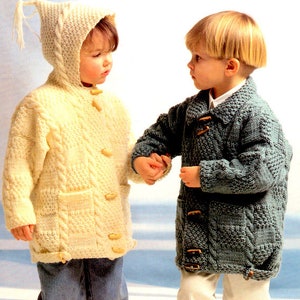 May include: Two children wearing knitted sweaters with hoods. The girl is wearing a cream sweater with a hood and a tassel. The boy is wearing a blue sweater with a hood. Both sweaters have a cable knit pattern and button closures.