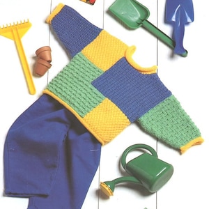 May include: A knitted baby sweater with a patchwork design in blue, green, and yellow. The sweater is laid out on a white surface with a pair of blue trousers, a rake, a watering can, and a small spade.