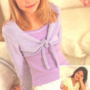 May include: A young girl wearing a lavender knitted shrug with a tie at the front. The shrug is worn over a lavender tank top and white skirt. The girl is sitting on a white couch. The image includes the text "Sirdar".