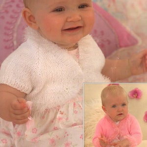 Op de afbeelding: Twee baby's dragen witte en roze gebreide vesten met een pluche rand. De vesten zijn open aan de voorkant en hebben een shawlkraag. De baby's dragen bloemenjurken.