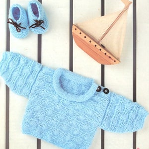 Può includere: Un maglione di lana blu chiaro per neonato con scollo rotondo e chiusura a bottoni. Il maglione è steso su una superficie a righe con una piccola barca di legno giocattolo.
