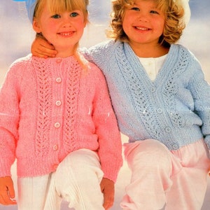 May include: Two young girls wearing knitted cardigans. The girl on the left is wearing a pink cardigan with a button-up front and a white collar. The girl on the right is wearing a light blue cardigan with a button-up front and a white collar.