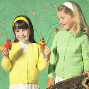 May include: Two young girls wearing knitted cardigans. The girl on the left is wearing a yellow cardigan and the girl on the right is wearing a green cardigan. Both girls are holding toy birds.
