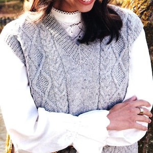 May include: A gray cable knit vest with a V-neck. The vest is worn over a white blouse with a lace trim.