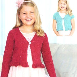 May include: A young girl wearing a red knitted cardigan with a ruffled hem and a single button closure. The cardigan has long sleeves and is paired with a light-colored dress. A smaller image shows a similar turquoise cardigan.