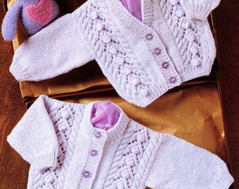 Peut inclure: Deux gilets en tricot pour bébé de couleur lavande avec des boutons. Les gilets ont un motif en tricot torsadé avec de petites boules.