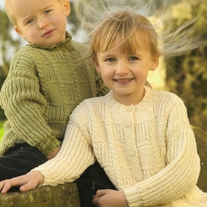 May include: Two children in hand-knitted sweaters. One wears a light green crew neck, the other a cream button-down cardigan. Both sweaters feature a textured, geometric pattern. The cardigan has button closures.