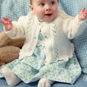 May include: A baby in a white knitted cardigan with decorative buttons, a blue floral dress, and white socks. The baby is sitting on a blue blanket. The cardigan has an open-knit design.